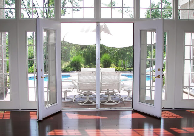 Open French doors reveal a sunny patio with a pool and outdoor seating.